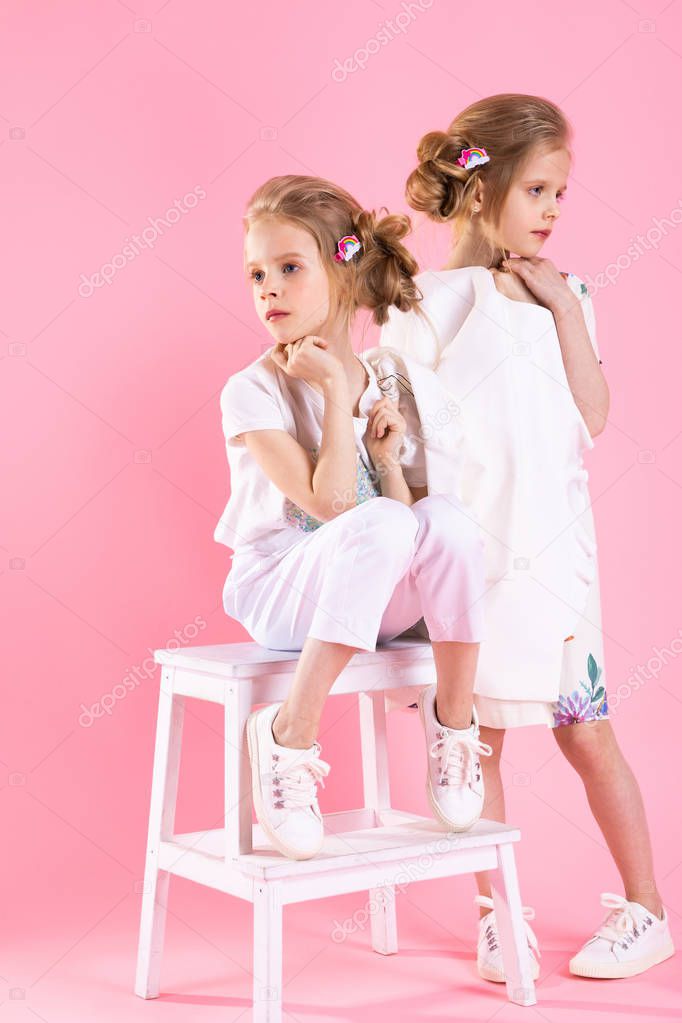 Twins girls in bright clothes posing near the stairs with two steps on a pink background.