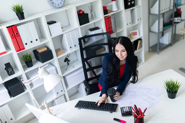 Bir bardak kahve ile onun elinde tutan ve tablo ofiste bir sandalyede oturan klavye, metin yazarak güzel genç kız.