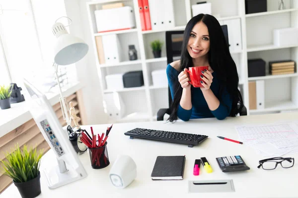 Mladá dívka sedí v kanceláři na stole a drží červený pohár ve svých rukou. — Stock fotografie