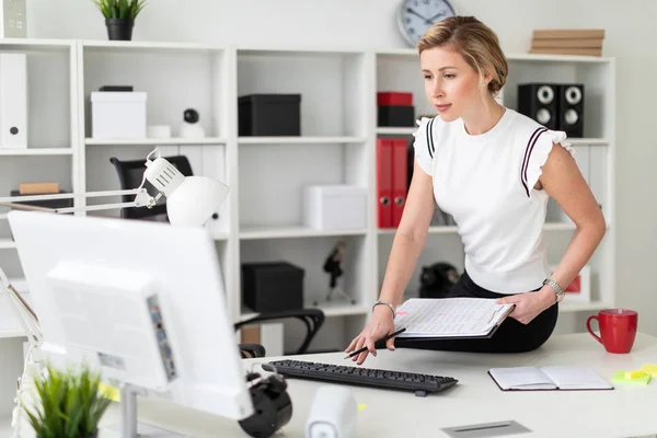 Młoda Blondynka siedzi na biurku w biurze, w ręku trzyma dokumenty i ołówek i drukuje na klawiaturze. — Zdjęcie stockowe