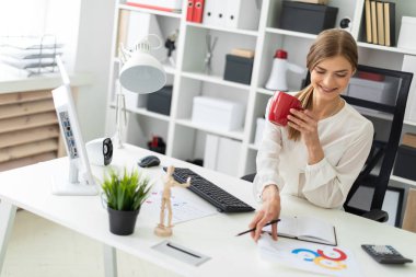 Genç bir kız ofiste bir masada oturuyor, kırmızı bir fincan elinde tutan ve bir levha ile bir grafik tablo alma.