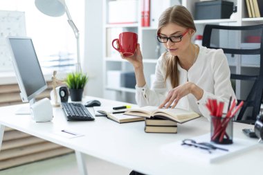 Genç bir kız ofiste bir masada oturur ve kırmızı bir fincan elinde tutan ve bir kitap okur.