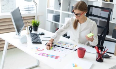 Genç bir kız ofiste bir masada oturuyor, elinde kalem tutan ve bir etiket sayfasında tutkallar.