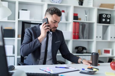 Bir adam ofiste masa başında oturan, telefonda konuşurken ve bir bardak kahve holding.