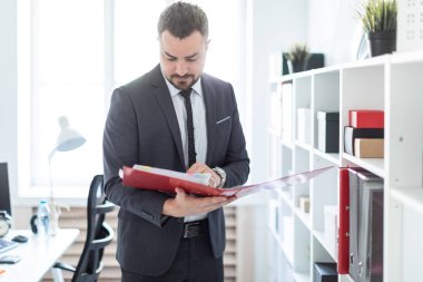 Adam raf ofiste yakınındaki duruyor ve bir klasör tutuyor.