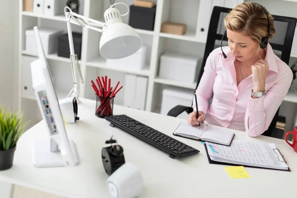 Молодая девушка в наушниках сидит за столом в офисе, держит карандаш в руке и работает с ноутбуком и компьютером . — стоковое фото
