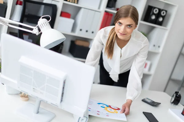 A young girl stands bent near a table in the office. The girl works with a computer, notepad and documents. — Stock Photo, Image