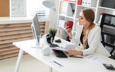 Genç bir kız ofiste bilgisayar masasında oturuyor ve bir fincan ve belgeleri tutuyor. Kız masada daha önce diyagram olan.