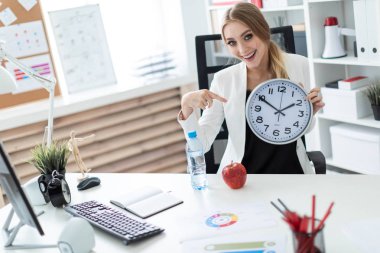 Genç bir kız ofiste bir masada oturur ve saat yüzünü puan bir parmak. Kız masada daha önce şurada bir elma ve bir şişe su.
