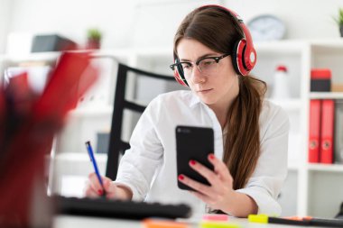 Genç bir kız kulaklık ofiste bir masada oturur ve bir kalem ve telefon elinde tutan.