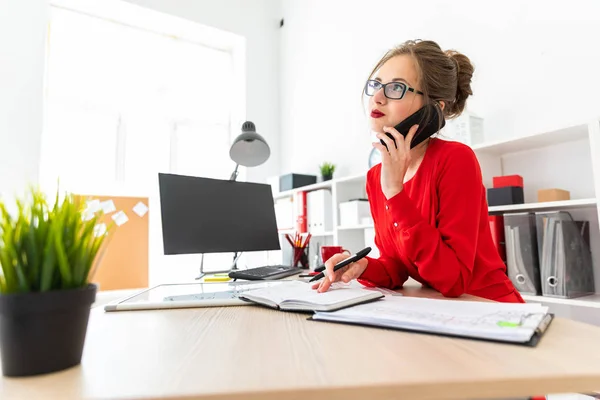 Een jong meisje is permanent aan een tafel in het kantoor, een zwarte marker houdt in haar hand en praten over de telefoon. Het meisje werkt met een computer, Kladblok en een magneetbord. — Stockfoto