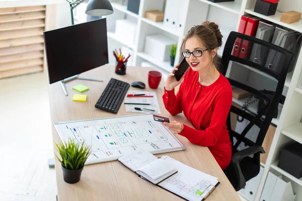 A young girl is sitting at the desk in the office, holding a bank card and phone in her hand. — Stock Photo, Image