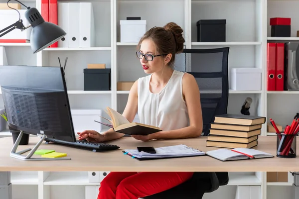 Uma jovem se senta em uma mesa de computador e segura um livro aberto em suas mãos. — Fotografia de Stock