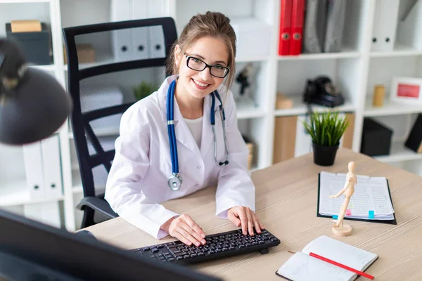 A young girl in a white robe is sitting at the computer desk and typing on the keyboard.