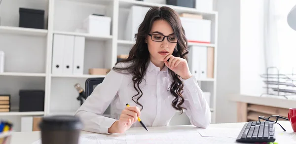 Una joven se sienta en la mesa de la oficina, sostiene un bolígrafo en la mano y mira los documentos . — Foto de Stock