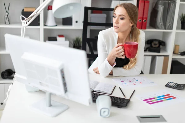 Una joven se sienta en un escritorio de computadora en la oficina y sostiene una taza roja en su mano. Antes de que la chica en la mesa son diagramas . —  Fotos de Stock