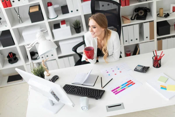 Молодая девушка сидит за компьютерным столом в офисе и держит чашку. Перед девушкой на столе диаграммы . — стоковое фото