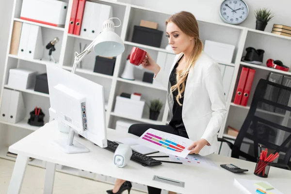 Девушка села на стол в своем кабинете, держа в руке красную чашку и работая с компьютером и документами . — стоковое фото