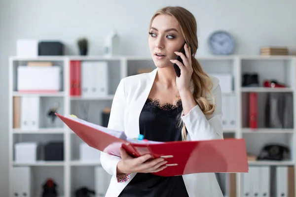 A young girl is standing in the office next to the shelving, talking on the phone and holding a folder with documents.