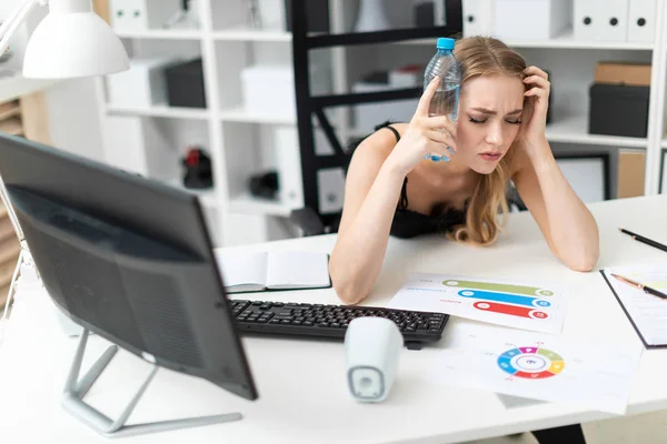 Ein junges Mädchen sitzt an einem Computertisch im Büro und hält sich eine Flasche Wasser an den Kopf. — Stockfoto