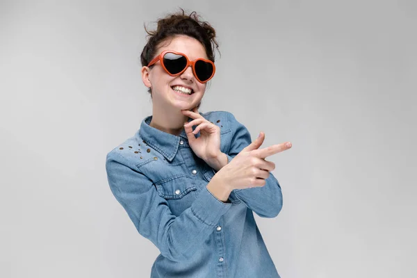 Junges brünettes Mädchen mit Brille in Form eines Herzens. Die Haare sind zu einem Dutt zusammengebunden. das Mädchen zeigt mit dem Finger. — Stockfoto