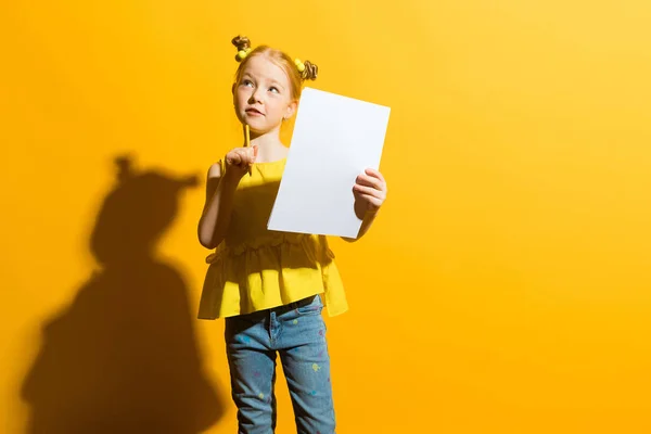 Mädchen mit roten Haaren auf gelbem Hintergrund. ein schönes Mädchen hält einen Bleistift und ein weißes Laken in ihren Händen. überlegte das Kind. — Stockfoto