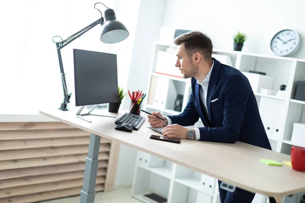 Ein junger Mann steht an einem Tisch im Büro, hält einen Bleistift in der Hand und arbeitet mit Dokumenten und einem Computer. — Stockfoto