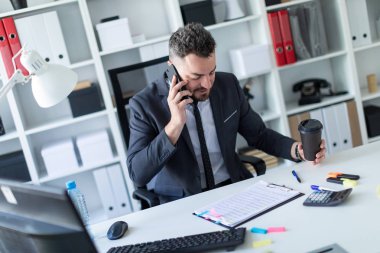 Bir adam ofiste masa başında oturan, telefonda konuşurken ve bir bardak kahve elinde tutan.