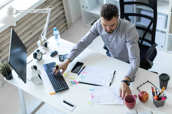Ofiste genç bir adam bir masada oturuyor, bir işaretleyici elinde tutuyor ve belgeler ve bir bilgisayar ile çalışır.