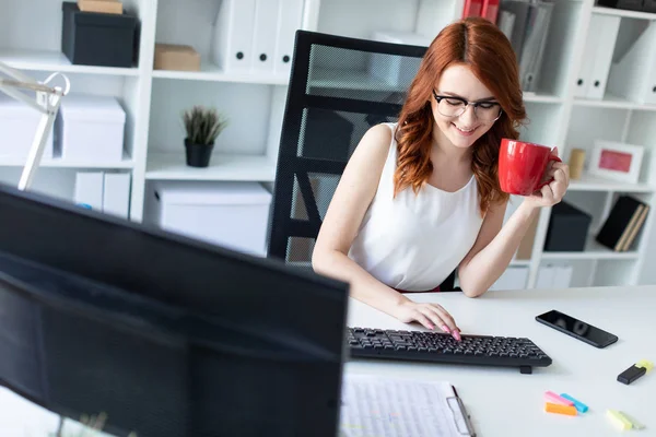 Güzel genç kız ofiste masada oturur, bir fincan bir elinde tutar ve bilgisayarı çalışır.