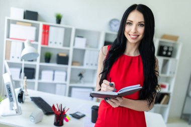 Kırmızı elbiseli güzel genç kız ofiste duran ve bir defter ve bir kalem tutan.