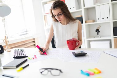 Ofisteki genç bir kız elinde pembe bir kalem, kırmızı bir kupa tutuyor ve belgelerle çalışıyor..