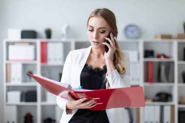 Genç bir kız ofis raf yanında duran, telefonda konuşurken ve belgeler ile bir klasör holding.