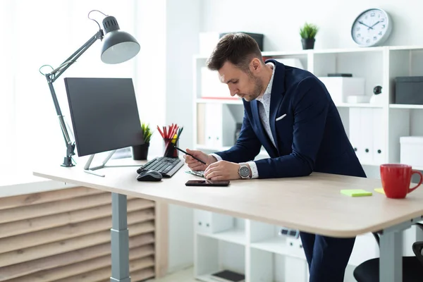 Молодой человек стоит возле стола в офисе, держит карандаш в руке и работает с документами и компьютером. . — стоковое фото