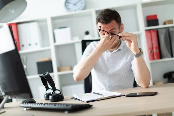 A young man sits at a table in the office, took off his glasses and rubbed his eyes. — Stock Photo, Image