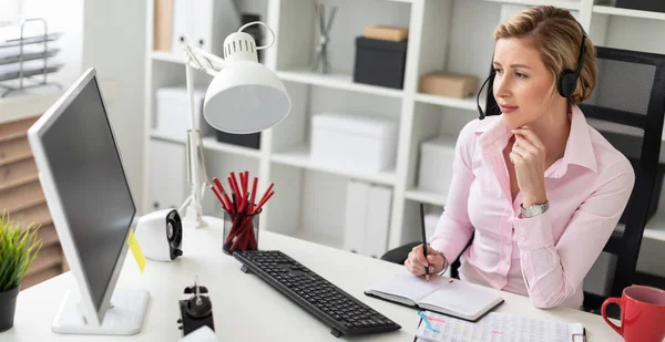 Une jeune fille en casque s'assoit à une table dans le bureau, tient un crayon dans sa main et travaille avec un cahier et un ordinateur . — Photo