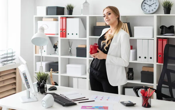 Genç bir kız ofis yakınındaki bir bilgisayar masası duruyor ve kırmızı bir fincan elinde tutan.