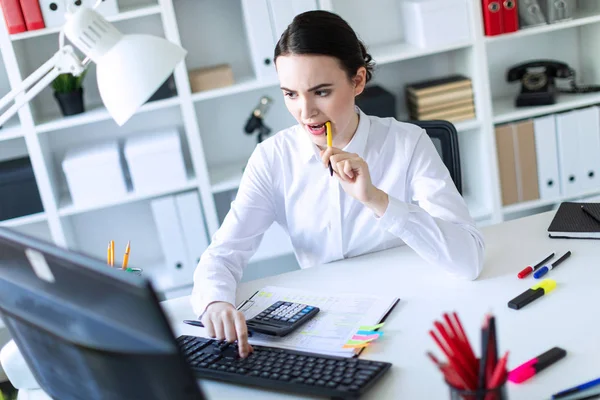 Een jong meisje in het Bureau houdt een pen in haar mond en werkt met een rekenmachine, documenten en een computer. — Stockfoto