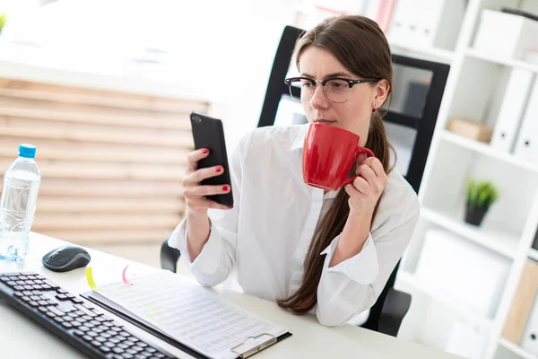 Genç bir kız bir telefon ve kırmızı bir fincan elinde tutan ofiste, bir masada oturuyor.