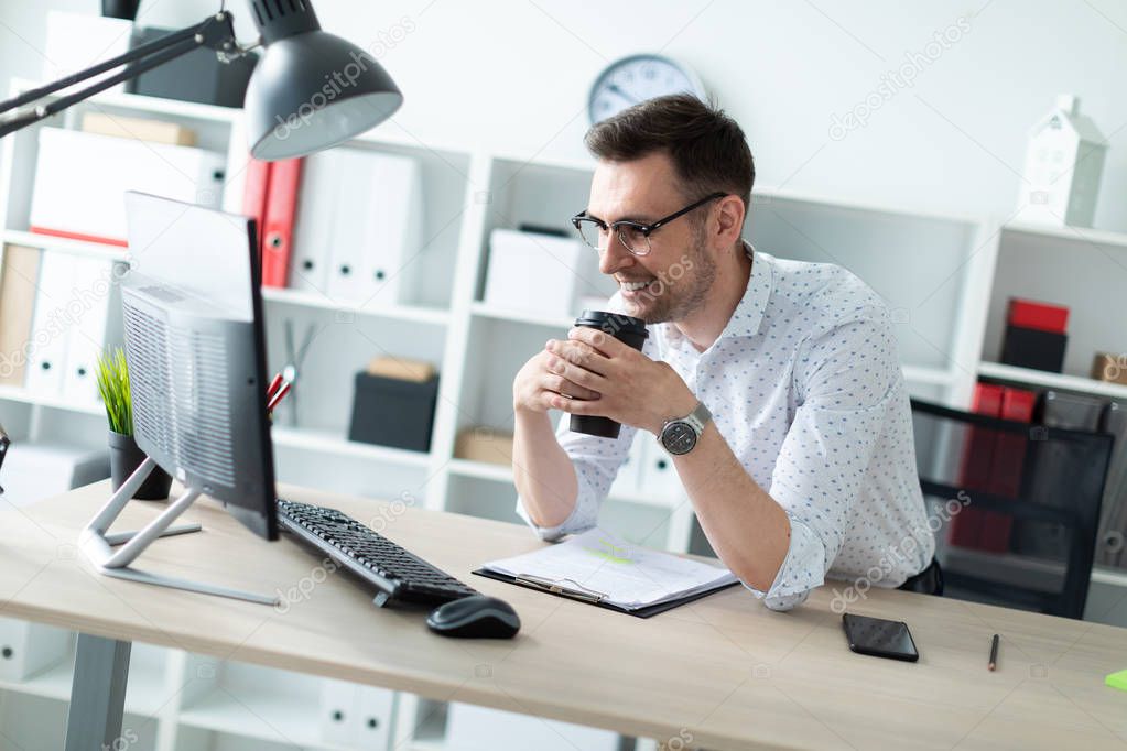 A young man in glasses stands near a table in the office holding a glass of coffee.