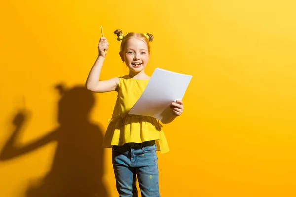 Girl with red hair on a yellow background. A beautiful girl is holding a pencil and a white sheet in her hands.