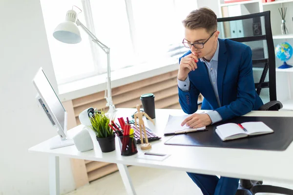 Genç bir adam ofiste bir masada oturuyor, kırmızı kalem elinde tutuyor ve belgeleriyle çalışır.