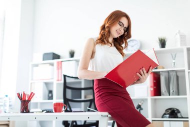 Güzel bir genç kız ofiste masanın üzerine oturdu ve elinde kırmızı bir dosya tutuyordu..