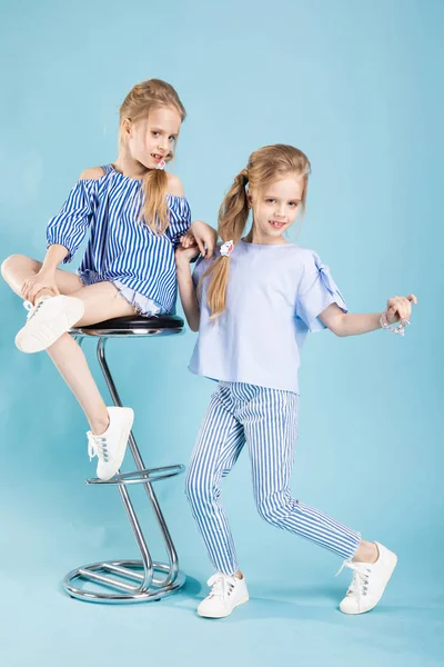 Bliźniaki dziewczyny w światło niebieskie stroje są pozowanie w pobliżu bar stołek na niebieskim tle. — Zdjęcie stockowe