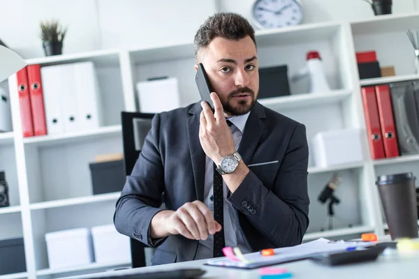 Bir adam ofiste, sol eliyle sağ kulak yakınındaki telefon tutarak masada oturuyor.