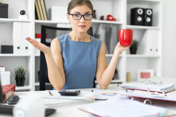 Молодая девушка сидит за столом в офисе, держа в руке красную чашку и работая с документами . — стоковое фото