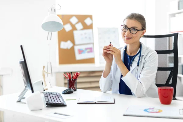 Ein junges Mädchen im weißen Gewand sitzt an den Tischen im Büro und hält einen Stift in der Hand. Ein Stethoskop hängt um ihren Hals. — Stockfoto