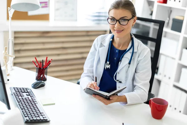 Una joven con una túnica blanca se sienta en una mesa en la oficina y sostiene un bolígrafo y un cuaderno en su mano. Un estetoscopio cuelga alrededor de su cuello . — Foto de Stock