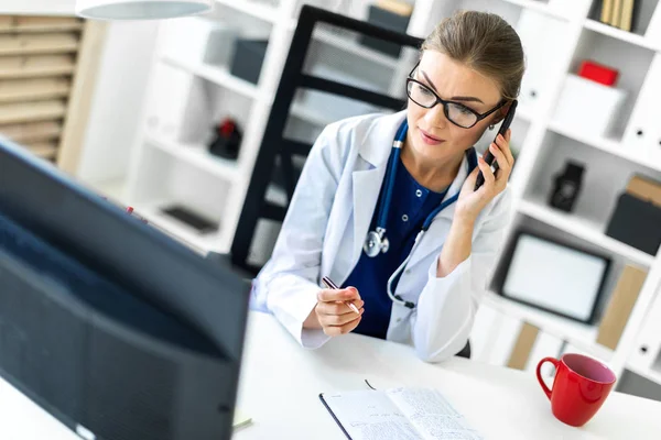 Una giovane ragazza vestita di bianco è seduta alla scrivania dell'ufficio, parla al telefono e tiene in mano una penna. Uno stetoscopio le pende intorno al collo . — Foto Stock