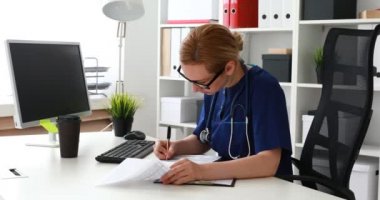 Genç bayan doktor masada dizüstü bilgisayarla ve belgelerle oturuyor ve formu dolduruyor. 